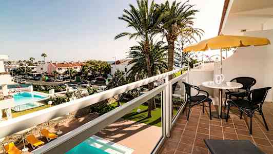 Utsikt fra en balkong med sittemøbler og parasoll, med et basseng og palmetrær i bakgrunnen. (AI-generert tekst)