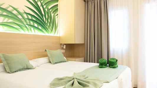 Et hotellrom med to enkeltsenger dekket av hvite laken og grønne dekorative puter. Veggdekorasjon med bladmotiv samt grønne sammenrullede håndklær på sengen. (AI-generert tekst)