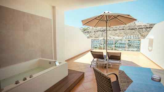 Terrasse med boblebadkar, parasoll, solsenger og utsikt over hus på en fjellside. (AI-generert tekst)