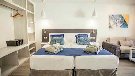 Dobbeltrom med to enkeltsenger, blått og hvitt sengetøy samt grå teppe. Moderne innredning med sofa og velbelyst atmosfære. (AI-generert tekst)