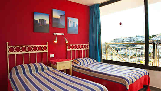 To enkeltsenger med stripete sengetøy og en rød vegg med bilder samt et stort vindu med utsikt over bygninger. (AI-generert tekst)