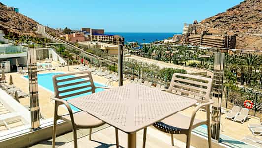 Utsikt over et basseng og solsenger med havet og bygninger i bakgrunnen, sett fra en balkong med bord og stoler. (AI-generert tekst)