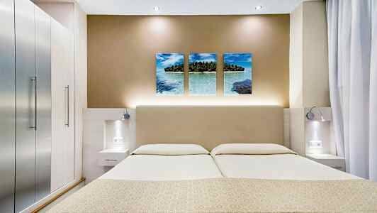 Bildet viser et moderne hotellrom med en dobbeltseng, stilrene sidebord og en veggdekor med tre bilder av et tropisk strandmotiv. (AI-generert tekst)