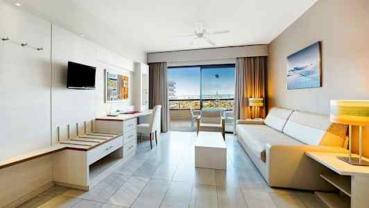 Lyst hotellrom med sofaer og møbler, åpent mot en balkong med havutsikt. (AI-generert tekst)