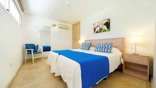 Et lyst hotellrom med en dobbeltseng, blå og hvite sengeklær, nattbord, veggdekorasjon og en lenestol. (AI-generert tekst)