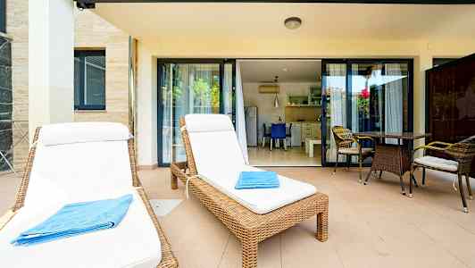 To solsenger med hvite puter og et blått håndkle på en terrasse med bord og stoler foran glassdører til et kjøkken. (AI-generert tekst)