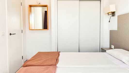 Et hotellrom med hvit innredning, en dobbeltseng med hvite laken og brune sengetepper, samt et speil på veggen. (AI-generert tekst)