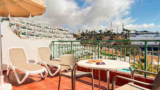 En terrasse med to solsenger, et bord og stoler samt utsikt over et bassengområde og palmer. (AI-generert tekst)