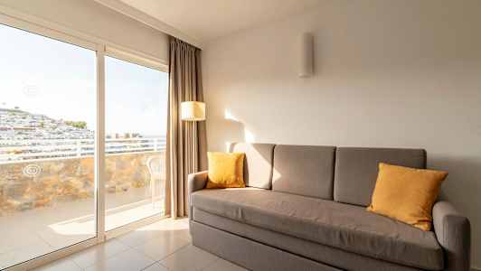 En grå sofa med gule puter står i et rom med store glassdører som leder ut til en balkong. (AI-generert tekst)