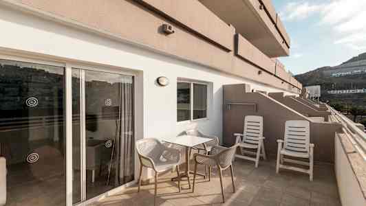 En terrasse med bord, stoler og solsenger, samt utsikt mot omkringliggende bygninger og fjell. (AI-generert tekst)