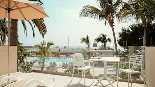 En balkong med solsenger og et bord med stoler, utsikt over et basseng og palmer. (AI-generert tekst)
