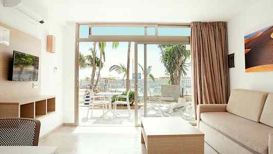 Et lyst rom med sofa, bord og TV samt utsikt mot en terrasse med bord og stoler. Palmer er synlige utendørs. (AI-generert tekst)