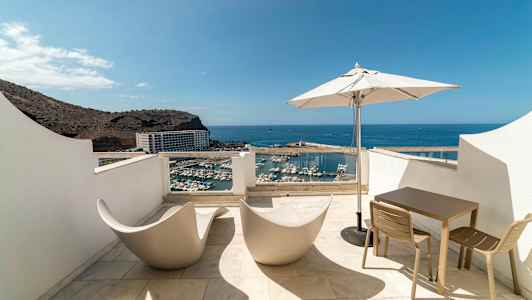 Terrasse med utsikt over havn og hav, utstyrt med moderne stoler, bord og parasoll. (AI-generert tekst)