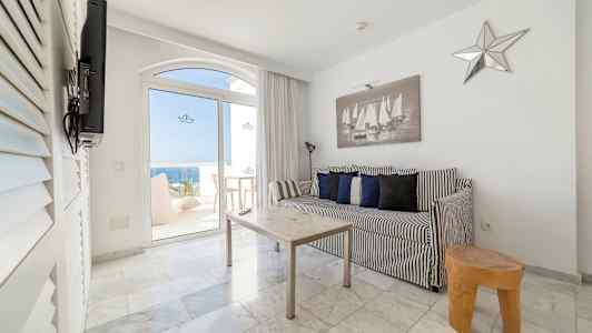 Svart-hvit interiør med sofa og bord i et rom som har utsikt gjennom glassdører mot sjøen. (AI-generert tekst)