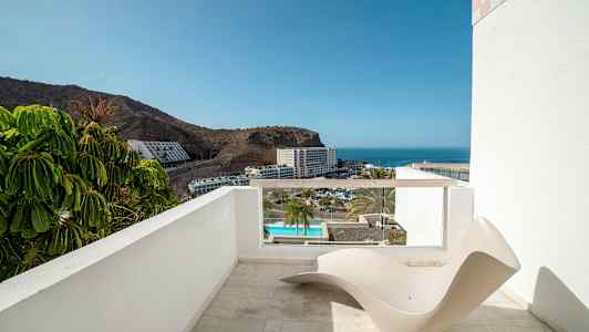 En terrasse med utsikt mot havet og et hotellområde, med en minimalistisk solstol i forgrunnen. (AI-generert tekst)