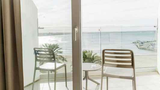 Utsikt fra en balkong med to stoler og et bord, mot en strand og havet. (AI-generert tekst)