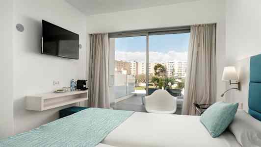 Et hotellrom med to senger oppredd med lyse laken og turkise sengetepp. En stol står foran en glassdør som leder ut til en balkong. En veggmontert TV henger ved siden av en hylle med vannflasker og kaffemaskin. (AI-generert tekst)