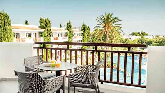 Utsikt fra en balkong med et bord og stoler der frokost er satt frem. I bakgrunnen er det et basseng, grøntområder og bygninger. (AI-generert tekst)