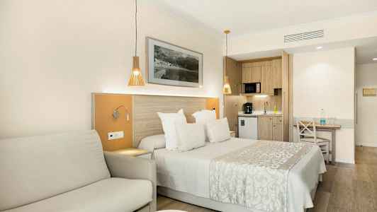 Et hotellrom med en dobbeltseng, sofa og et kjøkken i bakgrunnen. (AI-generert tekst)