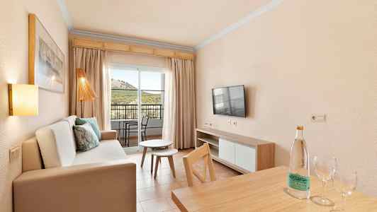 En stue med sofa, TV, bord og utsikt til en balkong med stoler. (AI-generert tekst)