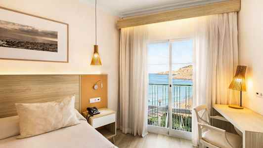 Et hotellrom med en seng og skrivebord, med utsikt mot havet gjennom et glassvindu med gardiner. (AI-generert tekst)