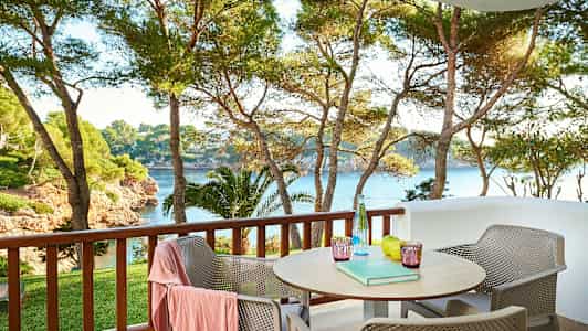 Utendørs veranda med bord og stoler, utsikt over trær og hav. (AI-generert tekst)