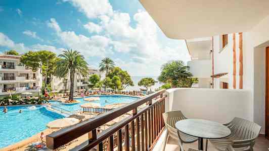 Utsikt fra en balkong mot et basseng omgitt av solsenger, palmetrær og bygninger. (AI-generert tekst)