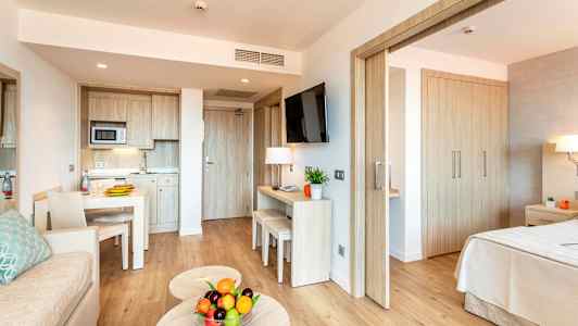 Moderne hotellrom med kjøkkenavdeling, bord, sofa og soverom. På sofabordet står en skål med fersk frukt. (AI-generert tekst)