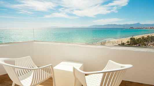 To hvite stoler og et bord på en balkong med utsikt over havet og stranden. (AI-generert tekst)