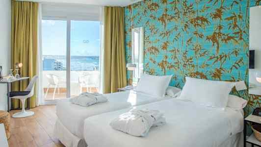 Et hotellrom med to hvite senger og balkongdør som fører til en terrasse med utsikt mot havet. (AI-generert tekst)