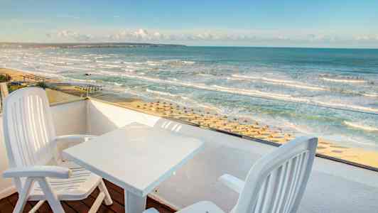 Utsikt fra en balkong med to stoler og et bord mot en strand med parasoller og bølgete hav. (AI-generert tekst)