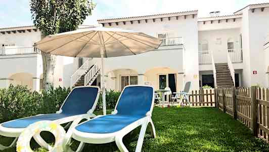 To solsenger med en parasoll på en gressplen foran en hvit bygning. (AI-generert tekst)