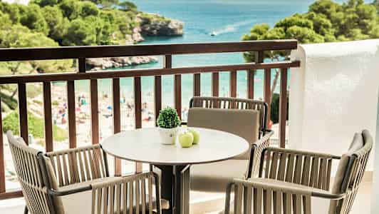 Balkong med bord og stoler som vender mot havet, stranden og frodige trær. (AI-generert tekst)