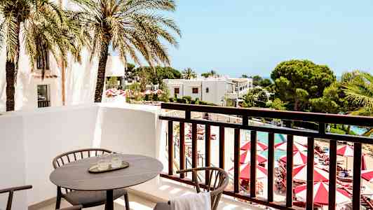 Utsikt fra en balkong med stoler og bord mot et basseng og omkringliggende hager. (AI-generert tekst)