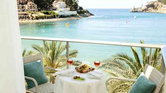 En balkong med bord og stoler, med utsikt over en bukt med palmer og bygninger i bakgrunnen. (AI-generert tekst)