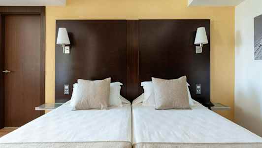 To enkeltsenger med hvite laken og beige puter mot en mørkebrun sengegavl, med vegglamper over. (AI-generert tekst)