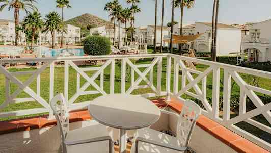 Utsikt fra en balkong med hvite utemøbler, mot en hage med basseng og palmer. (AI-generert tekst)