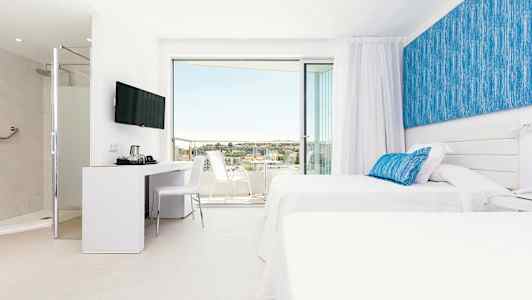 Et moderne hotellrom med to hvite senger og blå dekorasjoner. Rommet har en sittegruppe og en innglasset balkong med utsikt. (AI-generert tekst)