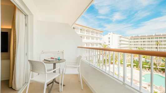 En møblert balkong med to stoler og et bord, med utsikt over hotellområdet og bassenget. (AI-generert tekst)