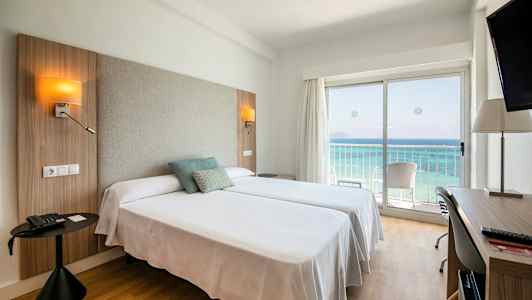 Hotellrom med to senger, havutsikt gjennom en glassdør og skrivebord. (AI-generert tekst)