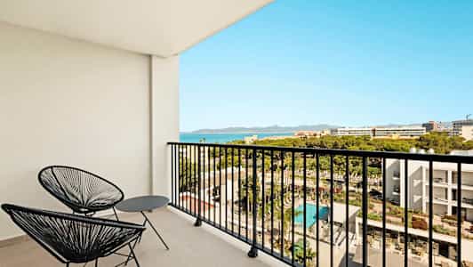 Balkong med to svarte stoler og bord, utsikt over hotellområde og hav. (AI-generert tekst)