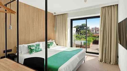 Et hotellrom med en seng oppredd med hvite laken og grønne puter, en stor glassdør som fører til en balkong med grøntområder. (AI-generert tekst)