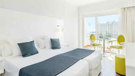 Et lyst hotellrom med to enkeltsenger, hvite møbler og en sittegruppe nær en glassdør som leder til en balkong. (AI-generert tekst)