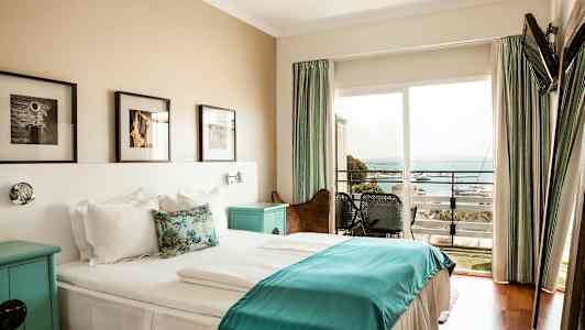 Et hotellrom med en dobbeltseng og sengetøy i hvitt og blått, veggkunst over sengen og en balkong med utsikt over havet. (AI-generert tekst)