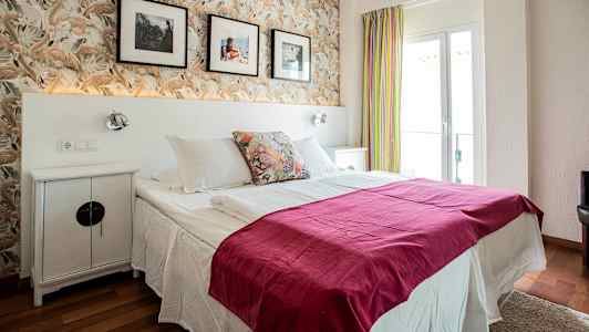 Et hotellrom med en dobbeltseng, pyntet med rosa sengeteppe og puter. Bak sengen er det en tapet med fjærmønster og veggdekorasjoner. (AI-generert tekst)