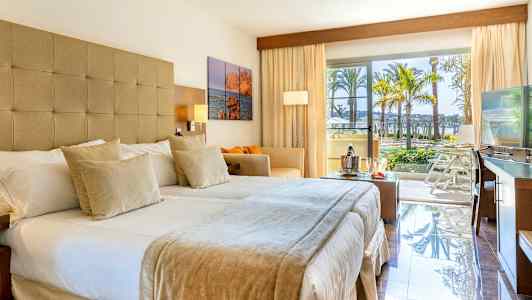 Hotellrom med dobbeltseng, sofa, TV og store vinduer som åpner ut mot en balkong med utsikt over palmer og bassengområde. (AI-generert tekst)