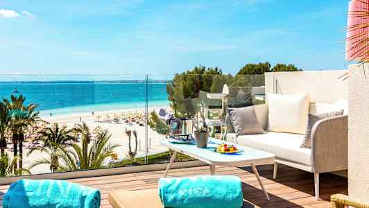 En terrasse med utsikt over en sandstrand, havet og parasoller, med en sofa, bord og håndklær i forgrunnen. (AI-generert tekst)