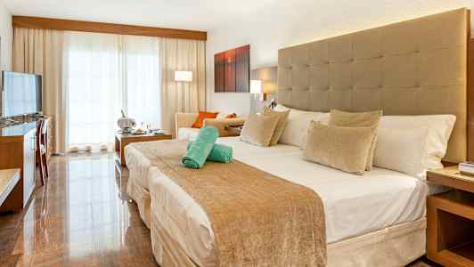 Hotellrom med en stor seng, sittegruppe og lyse gardiner foran en balkong. (AI-generert tekst)