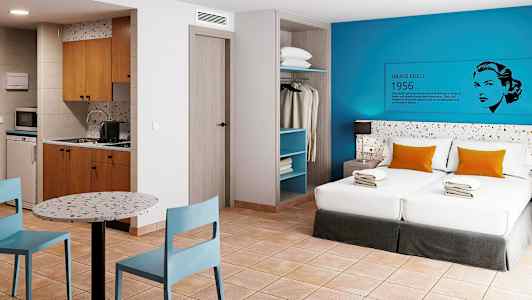 Et hotellrom med to senger, kjøkken, spisebord og åpen garderobe. (AI-generert tekst)