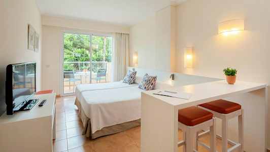 Et hotellrom med to enkeltsenger, en liten sittegruppe og en balkong med utsikt over grønne trær. (AI-generert tekst)